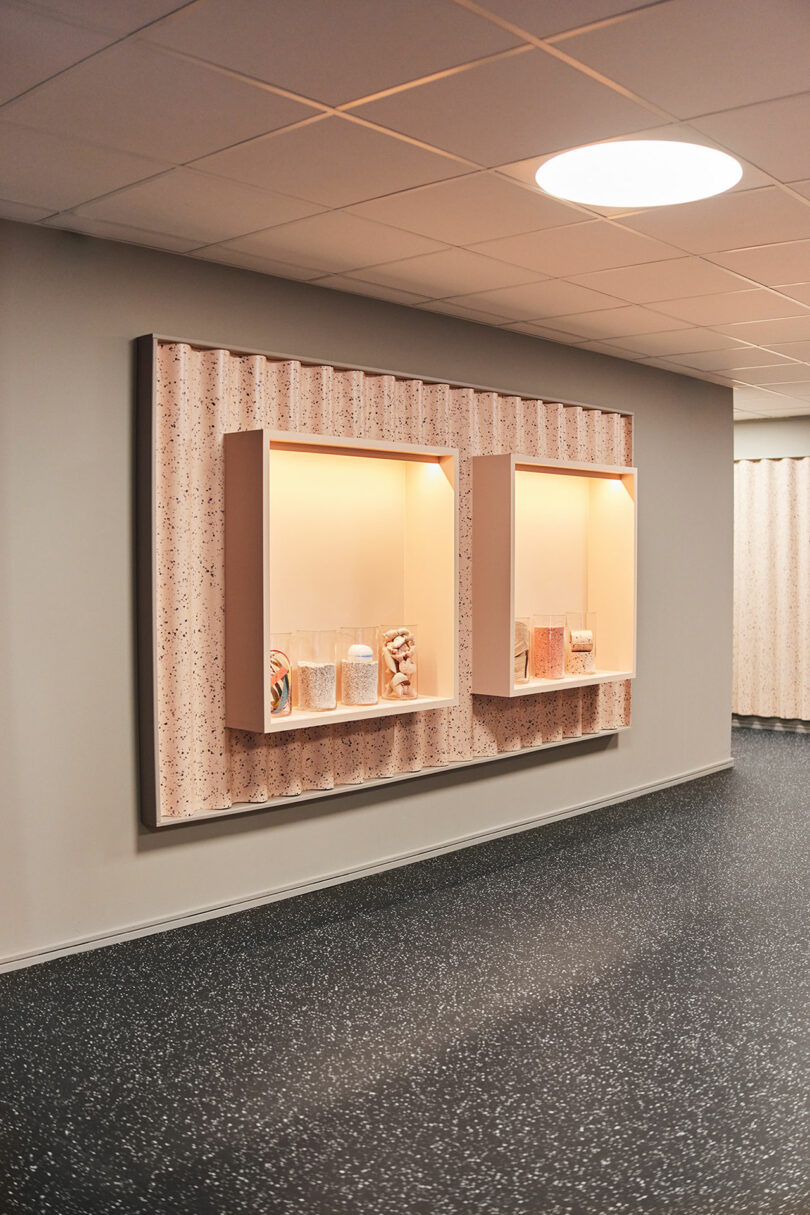 Two illuminated display cases mounted on a wall contain assorted objects and jars; the floor is dark speckled Tarkett, and the ceiling has square tiles with a round ceiling light.