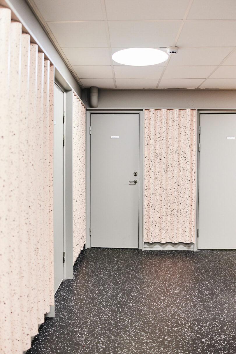 A hallway with speckled Tarkett partitions and closed gray doors, under a white ceiling with a round fluorescent light. The floor is dark with a speckled pattern.