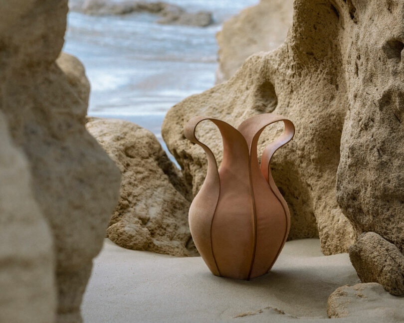 A sculptural, tan-colored ceramic vase with curved, petal-like edges sits on sandy ground among rocky formations near the water.
