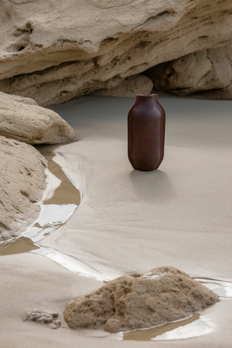 A dark brown vase stands upright on wet sand, surrounded by smooth rock formations and shallow water streams.