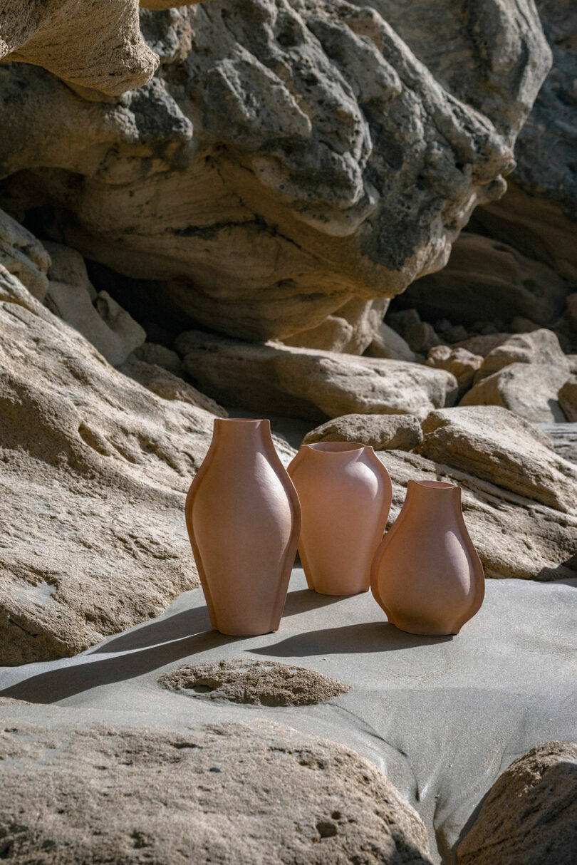 Three clay vases of varying sizes are placed on sandy ground among large, textured rock formations in natural sunlight.