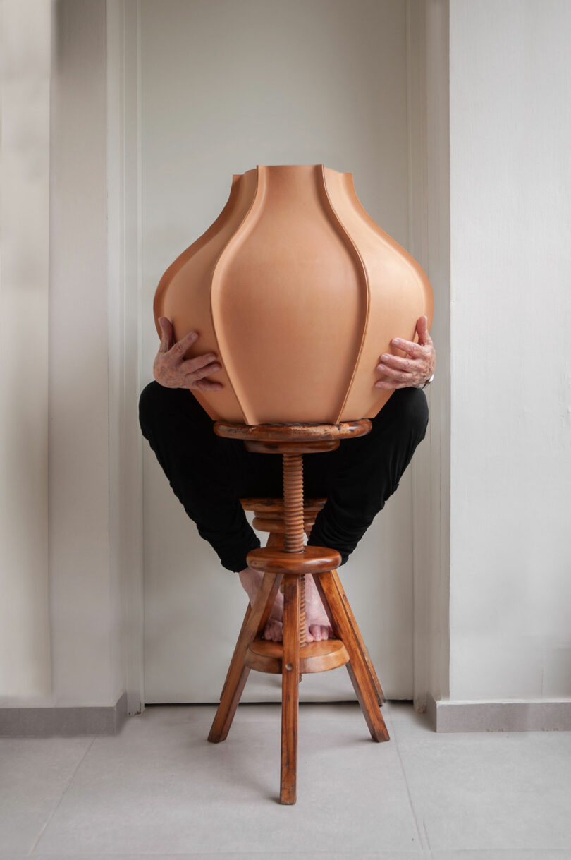 A person sitting on a wooden stool behind a large, round clay pot, with their hands gripping the sides and only their hands, feet, and legs visible.