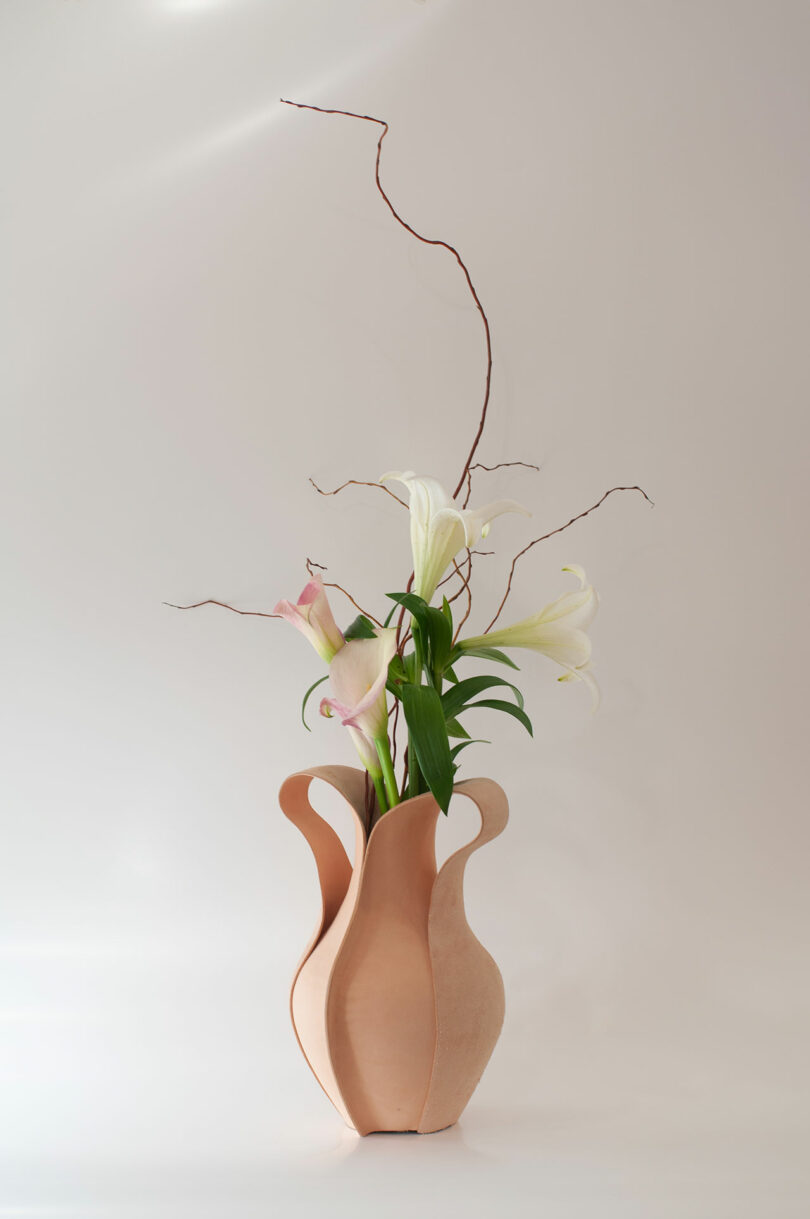 A sculpted beige vase holds white lilies, pink calla lilies, and long, twisting branches against a plain light background.