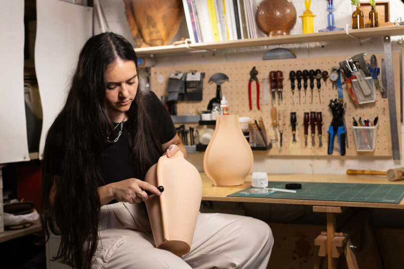 A person with long dark hair works on a ceramic vase in a workshop, surrounded by tools and materials on a wall-mounted pegboard.