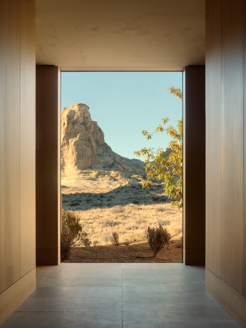 A large window in a modern wood paneled hallway frames a view of a rocky desert landscape with dry grass, bushes and a clear sky.