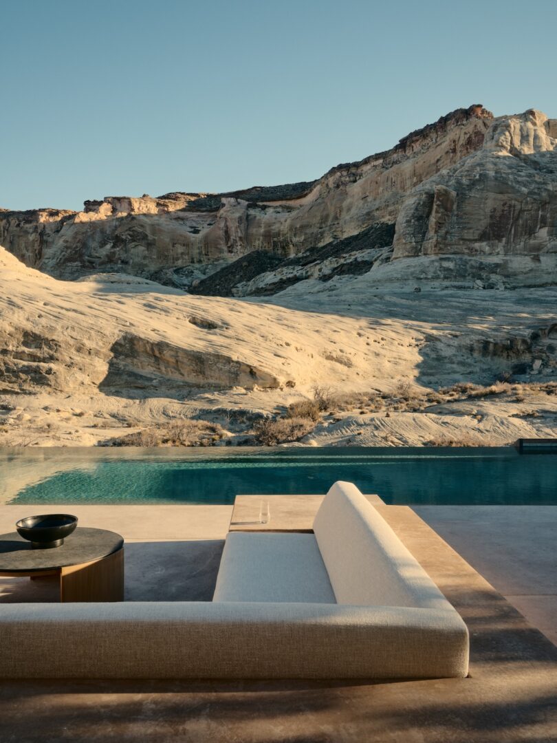 A modern outdoor sofa and table sits next to a pool overlooking the rocky desert cliffs under a clear sky.