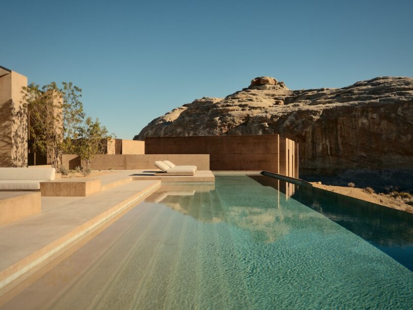 Modern infinity pool with sunbeds, against the backdrop of desert architecture and rocky landscape under a clear blue sky.