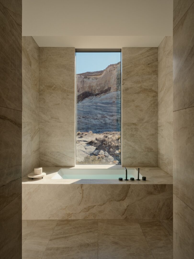 Modern bathroom with beige stone walls and floors, with a built-in bathtub under a tall window framing the view of a rocky desert landscape.