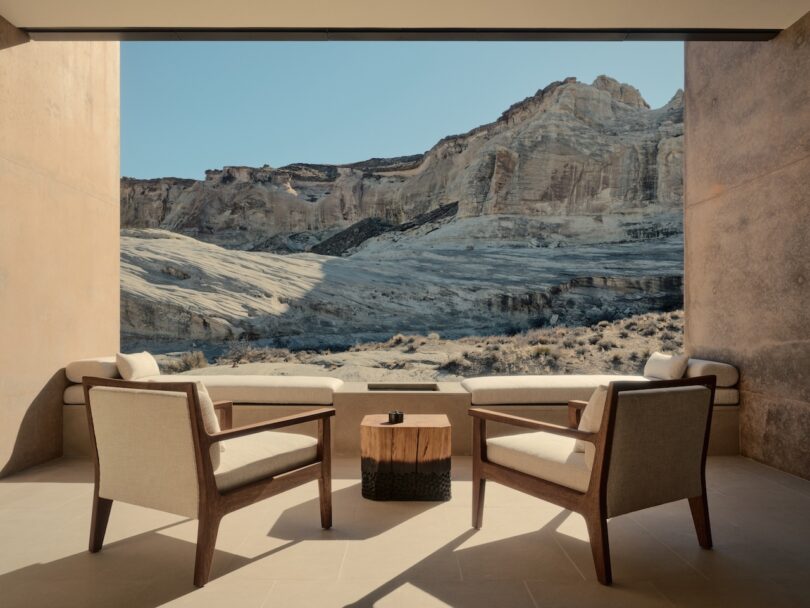 Two modern armchairs and a small table on a patio overlook a rocky desert landscape with tall beige cliffs under a clear blue sky.