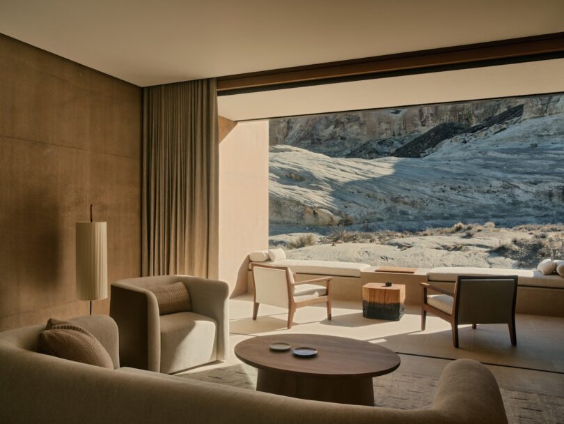 A modern neutral-toned living room with large windows opens to a rocky desert landscape. Minimal furniture includes armchairs, a sofa and a round table.