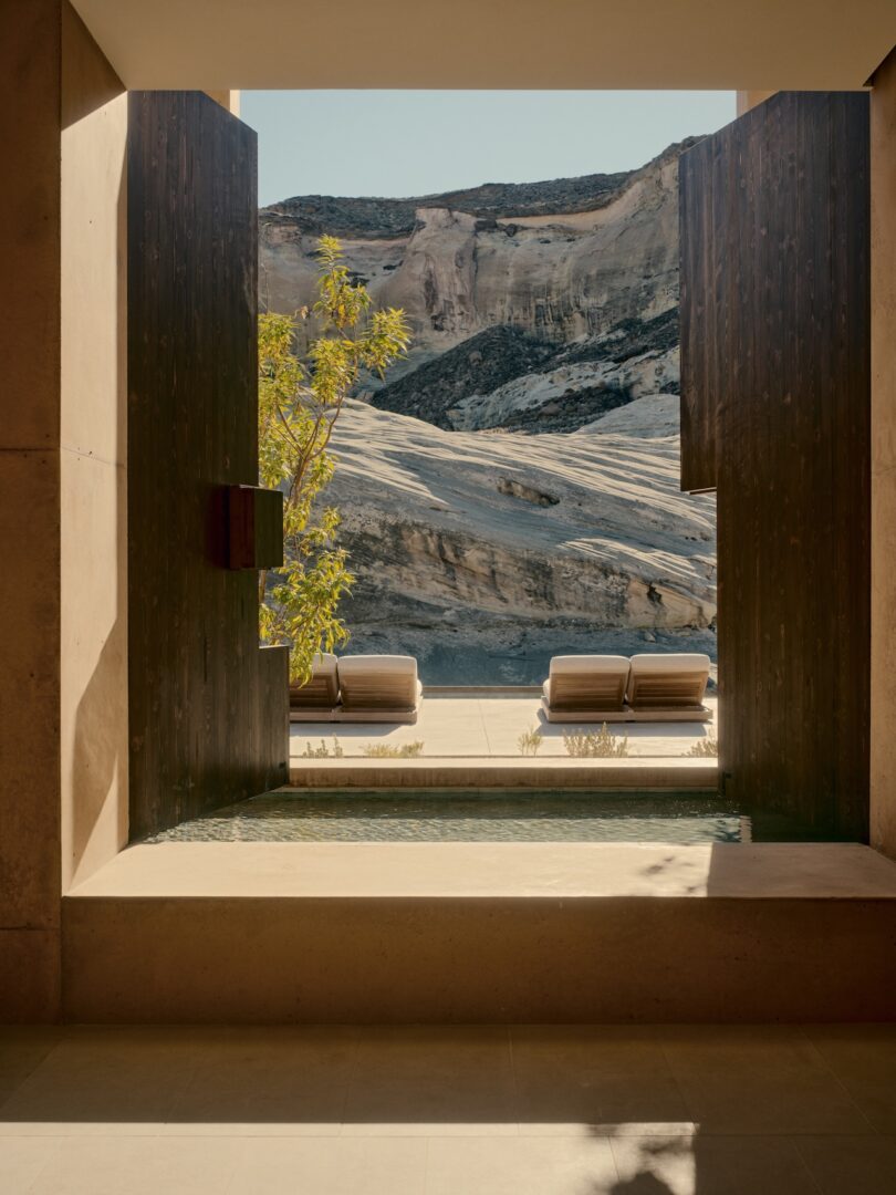 View through a modern stone and wood opening to a small pool, sunbeds and rocky desert cliffs in the background.