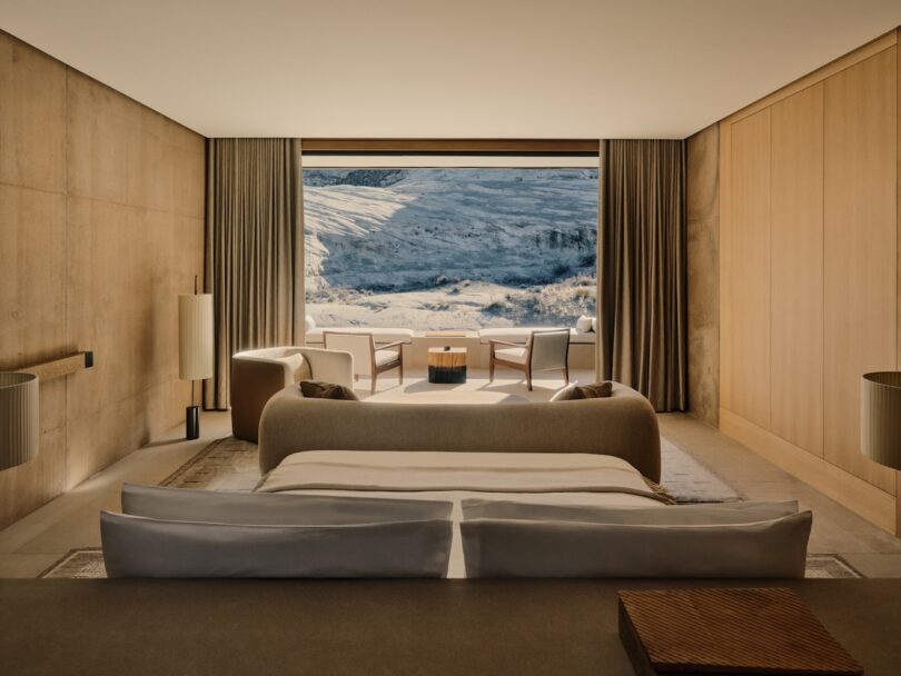Modern, minimalist bedroom with neutral tones, large bed and sitting area, with floor-to-ceiling windows overlooking a snowy mountain landscape.