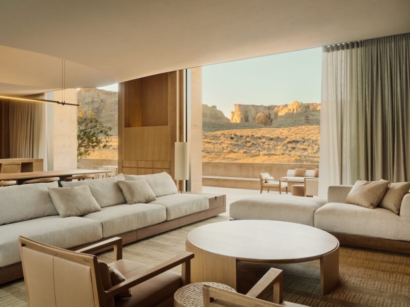 Modern living room with furniture in neutral tones, large windows and views of a desert landscape with rock formations outside.
