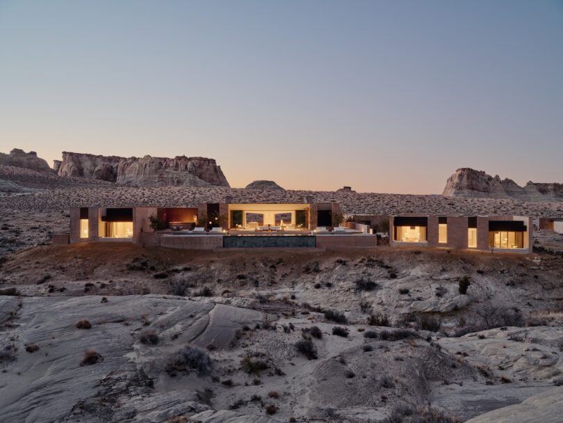 A modern flat-roofed building with large glass windows is illuminated at dusk, against a rocky desert landscape with midges in the background.