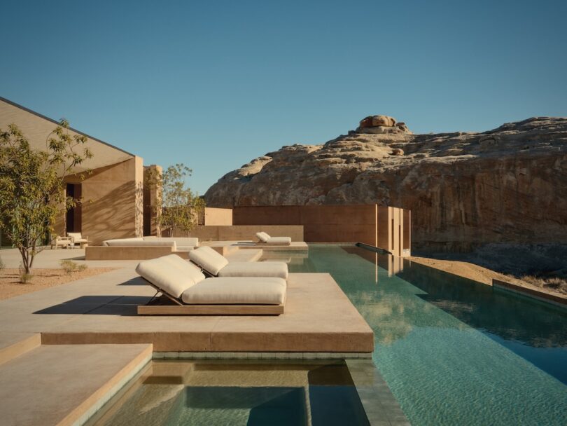 Modern outdoor swimming pool with sunbeds next to a minimalist building, against the backdrop of a rocky desert landscape under a clear blue sky.