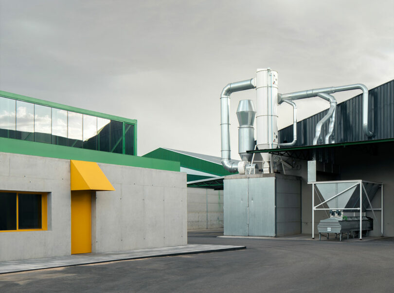 Una instalación industrial con edificios de hormigón, puertas y ventanas de color amarillo brillante, un techo de metal verde y grandes conductos de ventilación plateados contra un cielo nublado es el hogar del innovador Impepinable Studio.