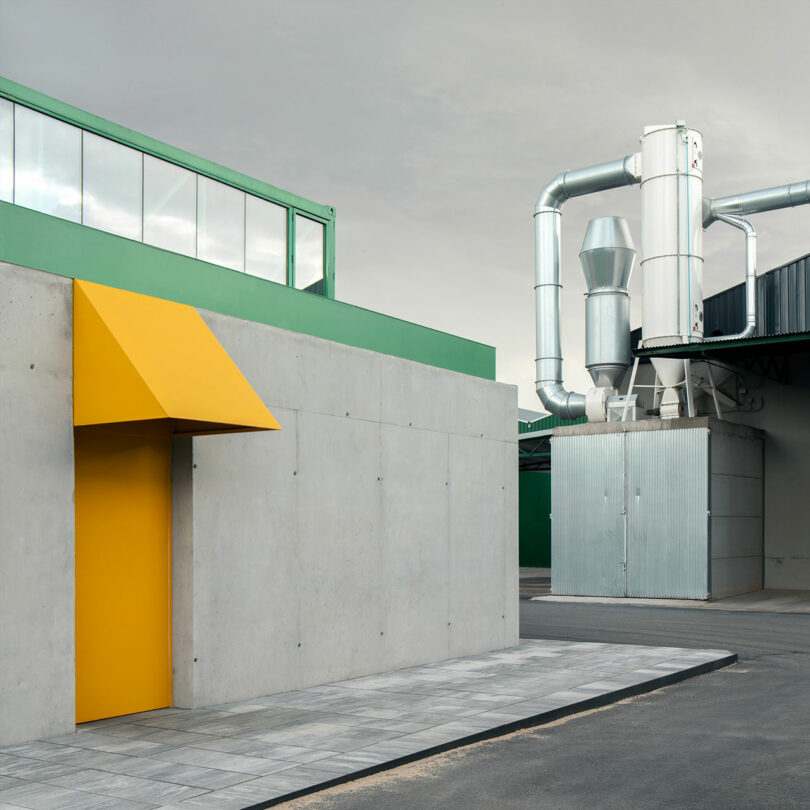 Un moderno edificio industrial que podría ser un insoportable estudio, con paredes de hormigón gris, puertas de color amarillo brillante, conductos de ventilación metálicos y detalles en verde contra un cielo nublado.