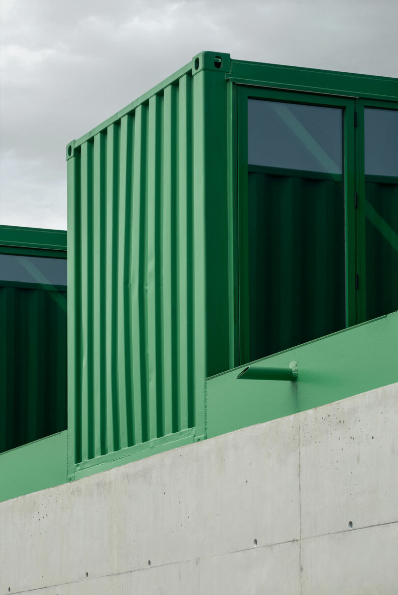 La estructura verde del contenedor de envío de Impepinable Studio se asienta sobre un muro de concreto con un cielo nublado de fondo.