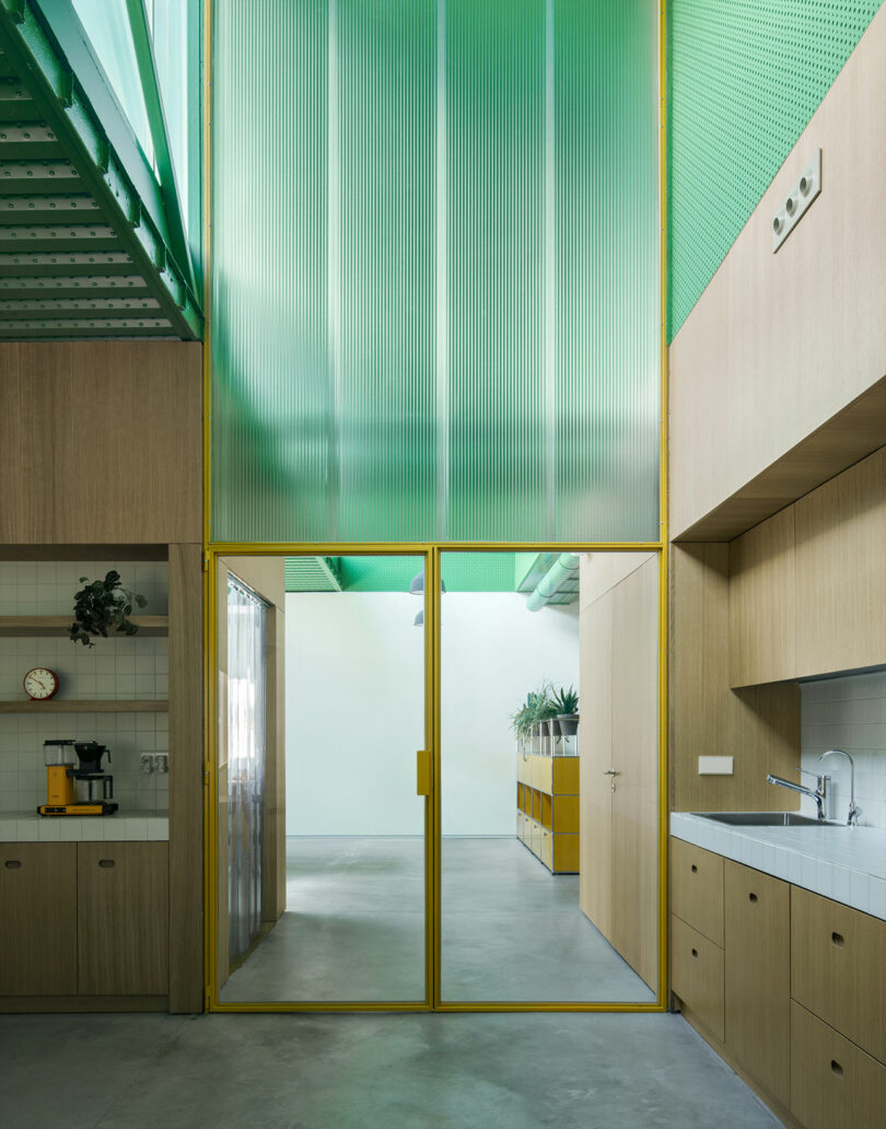 Impepinable Studio diseñó una cocina moderna con gabinetes de madera, un protector contra salpicaduras de azulejos, una máquina de café y una puerta de vidrio con marco amarillo que se abre a un pasillo con un techo verde translúcido y piso de concreto.