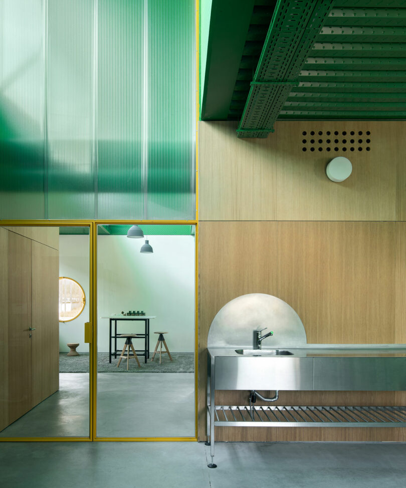 Impepinable Studio diseñó una cocina contemporánea con fregadero de acero inoxidable, paneles de madera, puertas de vidrio con marco amarillo y vigas verdes en el techo que fluye a la perfección hacia una habitación contigua minimalista.