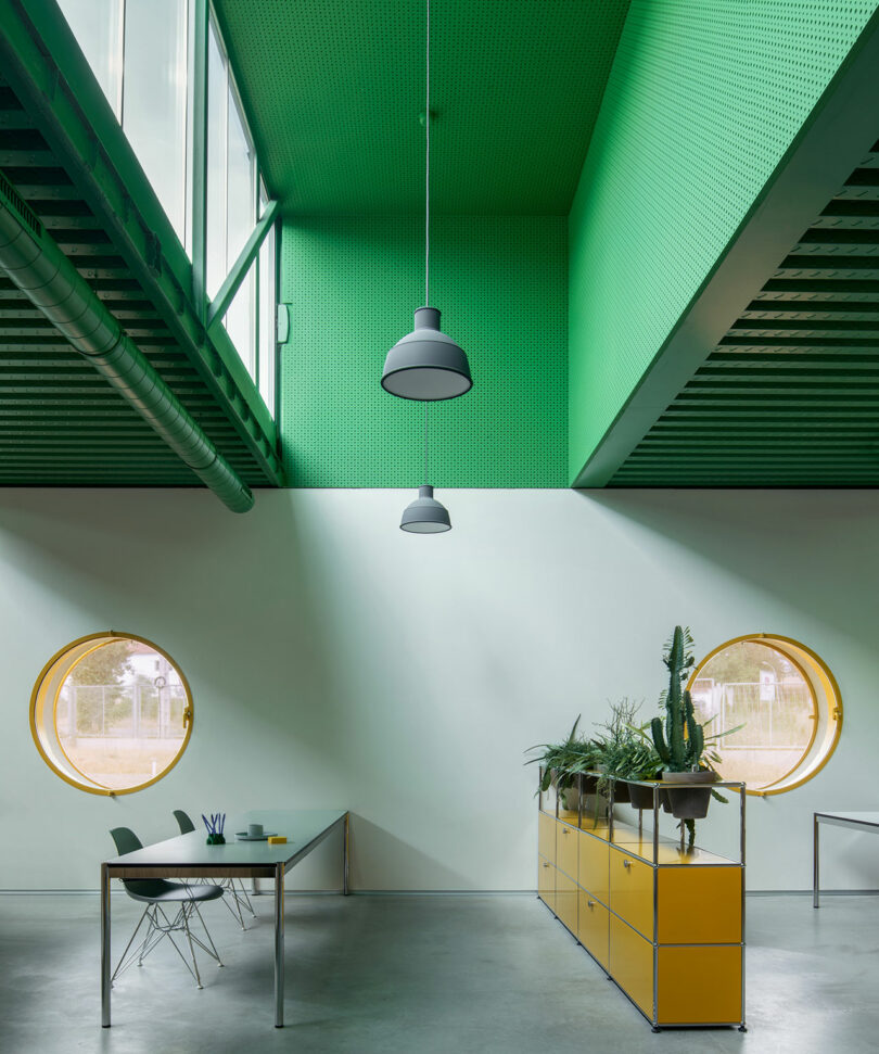 Impepinable Studio diseñó un espacio de oficina moderno con un techo verde, ventanas redondas amarillas, unidades de almacenamiento amarillas con plantas y una mesa minimalista con dos sillas.