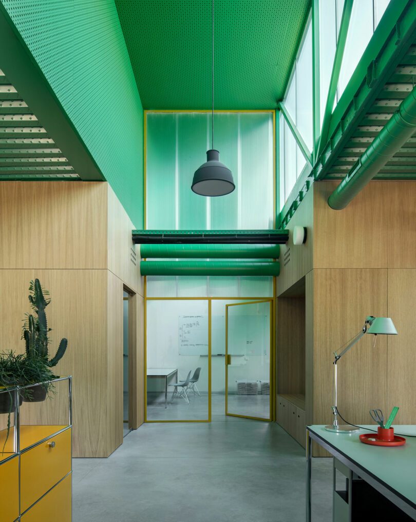 Interior de oficina moderno diseñado por Impepinable Studio con techos altos de color verde, paredes con paneles de madera, puertas de vidrio con marcos amarillos y muebles minimalistas que incluyen lámparas de mesa y plantas de cactus.