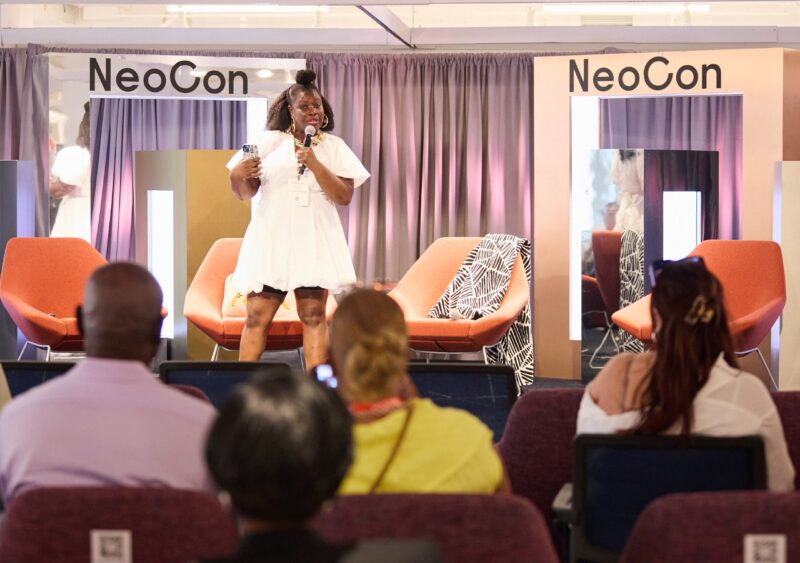 A woman in a white dress speaks on stage at 2026 NeoCon Talks, standing in front of modern chairs, while an audience listens.