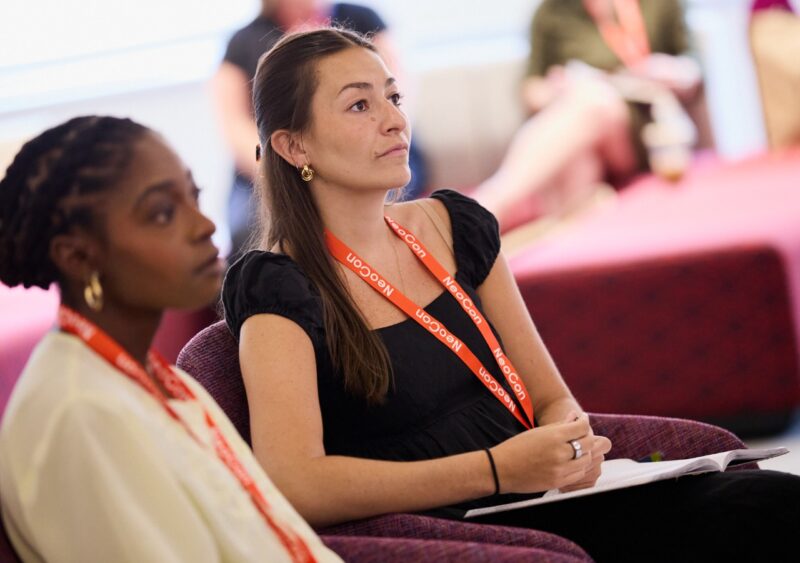 Two women wearing conference lanyards sit and listen attentively at the NeoCon Talks, one holding a notebook, in a brightly lit indoor setting with others in the background.