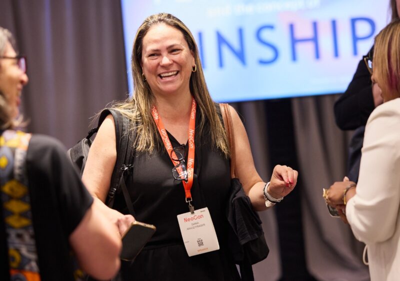 A woman wearing a NeoCon Talks badge smiles and converses with others at an indoor event, with a large screen in the background.