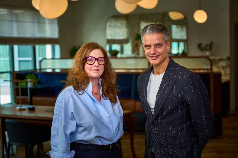 A woman in a blue shirt and glasses stands next to a man in a dark patterned suit in a modern office with round lights and plants in the background.