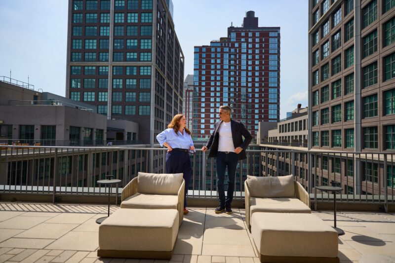 Two people stand and talk on a rooftop terrace between tall urban buildings, with two beige lounge chairs in the foreground.