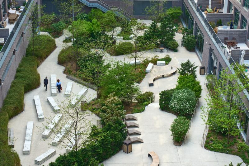 Aerial view of a modern urban courtyard with winding paths, benches, various green plants, and several people walking or sitting.