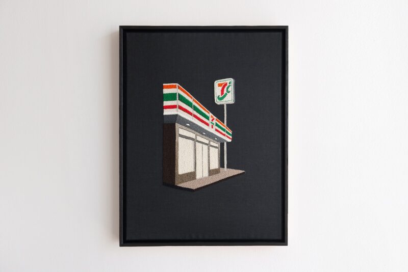 Framed embroidery artwork depicting a 7-Eleven convenience store and its sign against a black background, hung on a white wall.