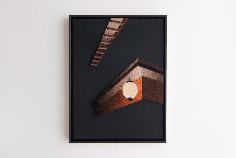 Framed textile artwork depicting geometric roof shapes and a white lantern on a dark background, hanging on a white wall.