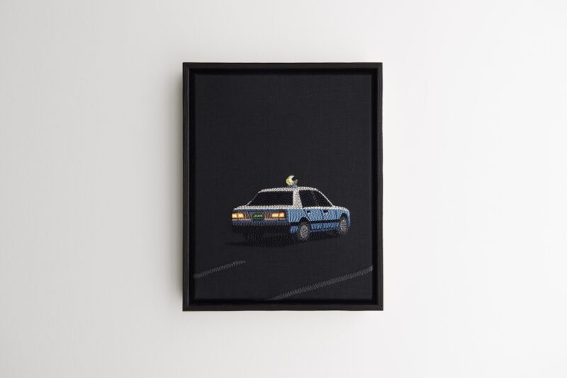 Framed embroidery art of a blue and white taxi with a yellow sign on top, depicted driving on a dark background, hanging on a white wall.