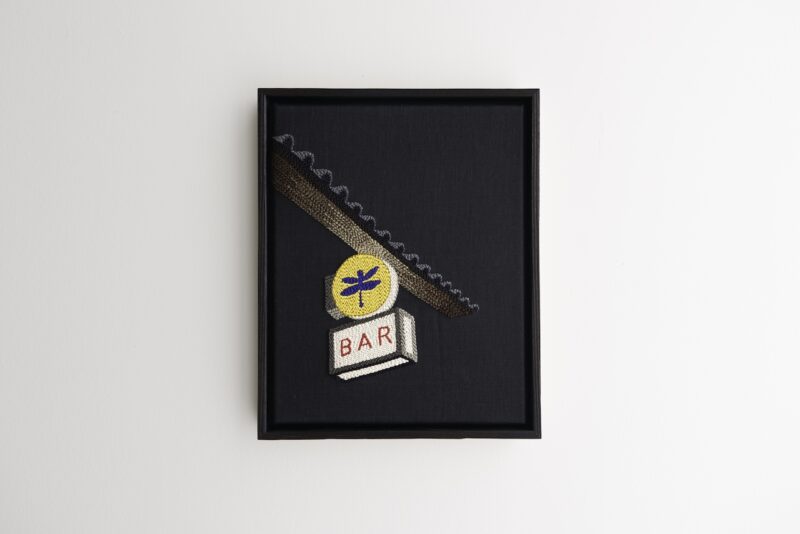 Framed embroidery of a bar sign with a dragonfly emblem and the word "BAR" on a dark background, hanging on a white wall.