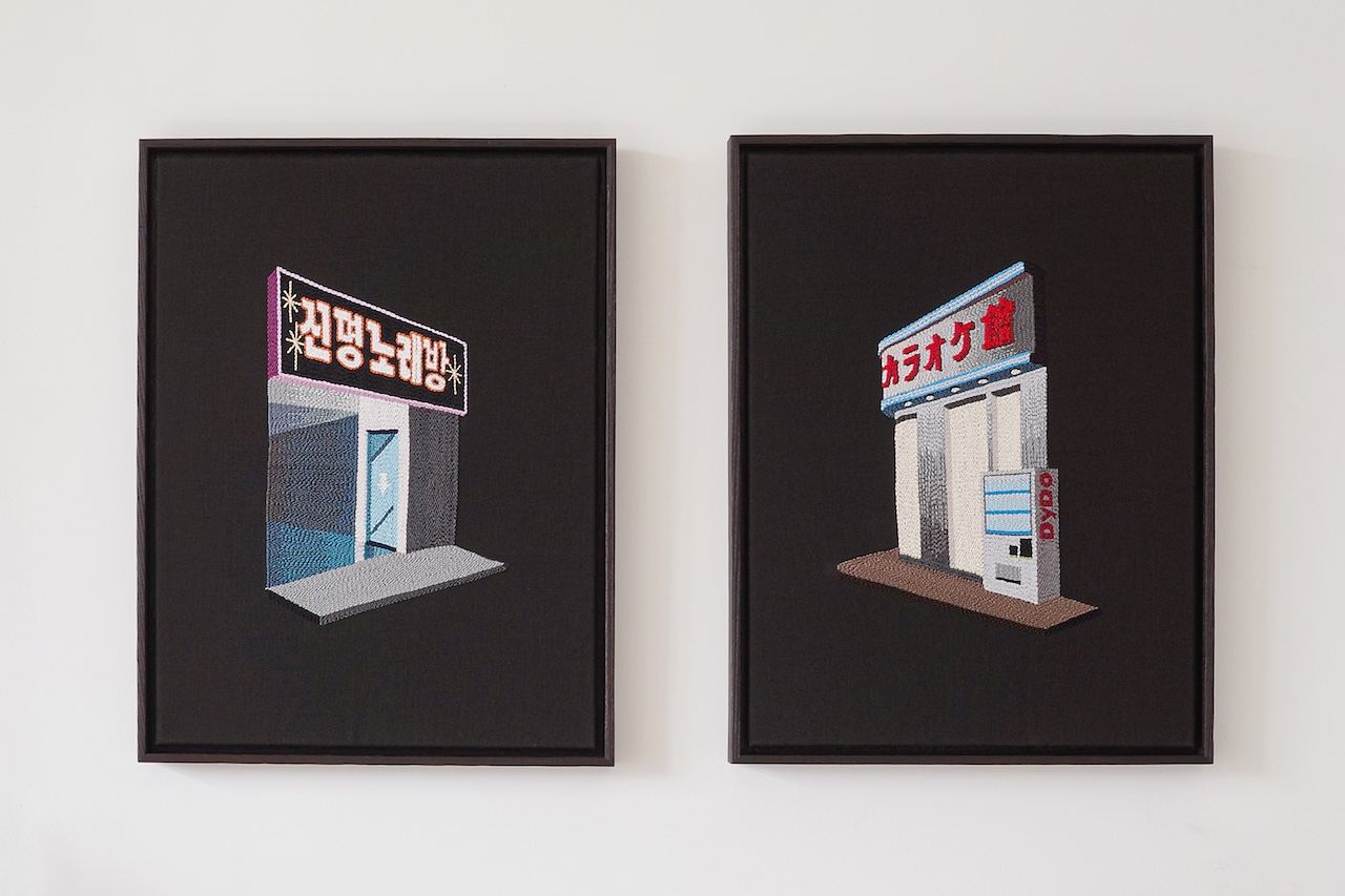 Two framed artworks depict stylized storefronts with foreign script signage, displayed side by side on a plain white wall.