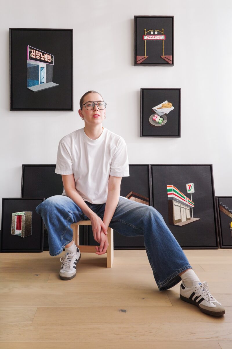 A person in a white t-shirt and jeans sits on a stool in front of a wall displaying framed artworks, some depicting storefronts and signs.