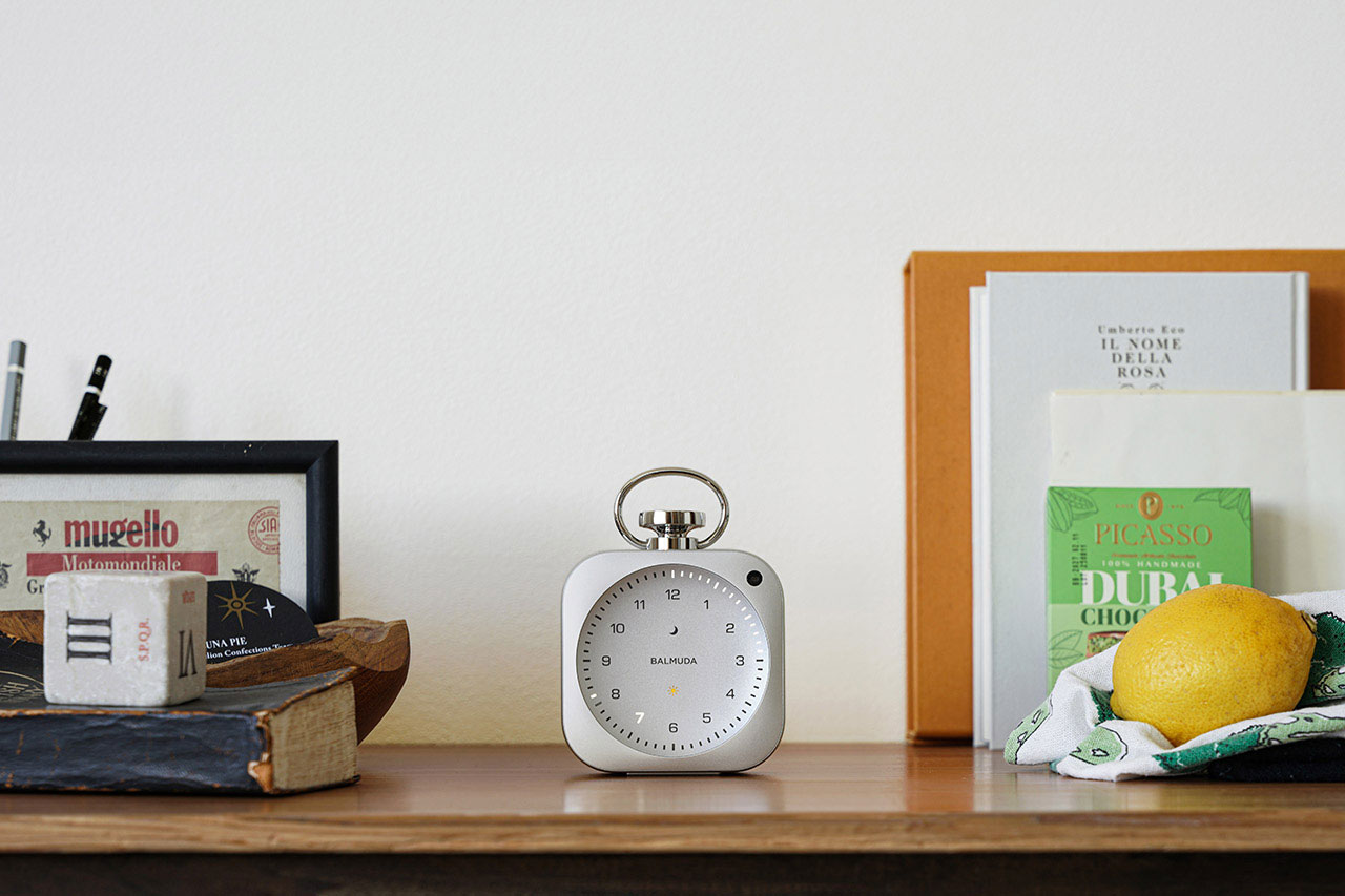 A silver alarm clock sits on a wooden shelf surrounded by books, a framed picture, pens, and a lemon on a cloth.