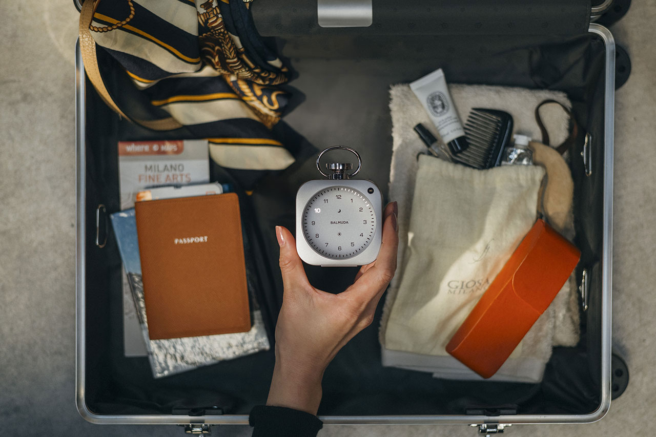 A hand holds a small clock over an open suitcase packed with a passport, books, a scarf, a brush, an eyeglass case, and other travel essentials.