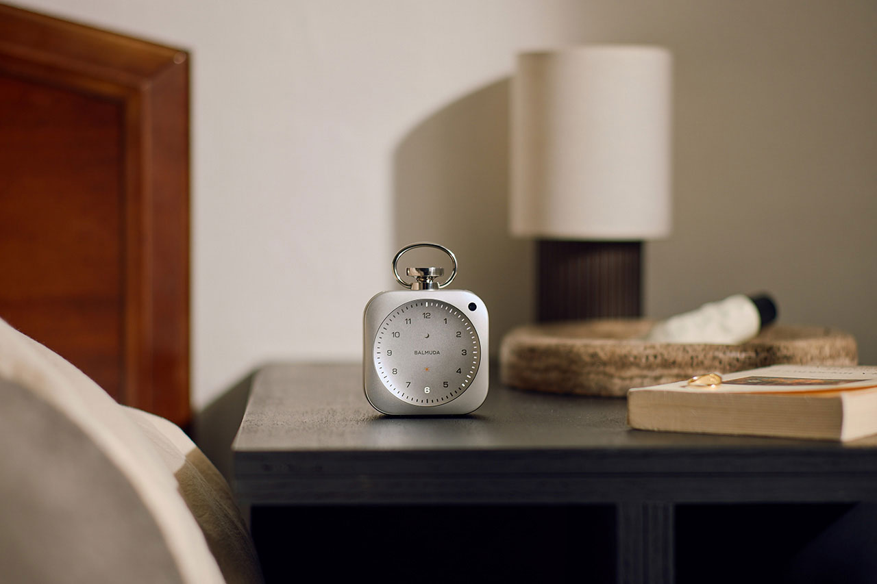 A silver alarm clock sits on a bedside table next to a lamp, a woven tray with a small item, and an open book.