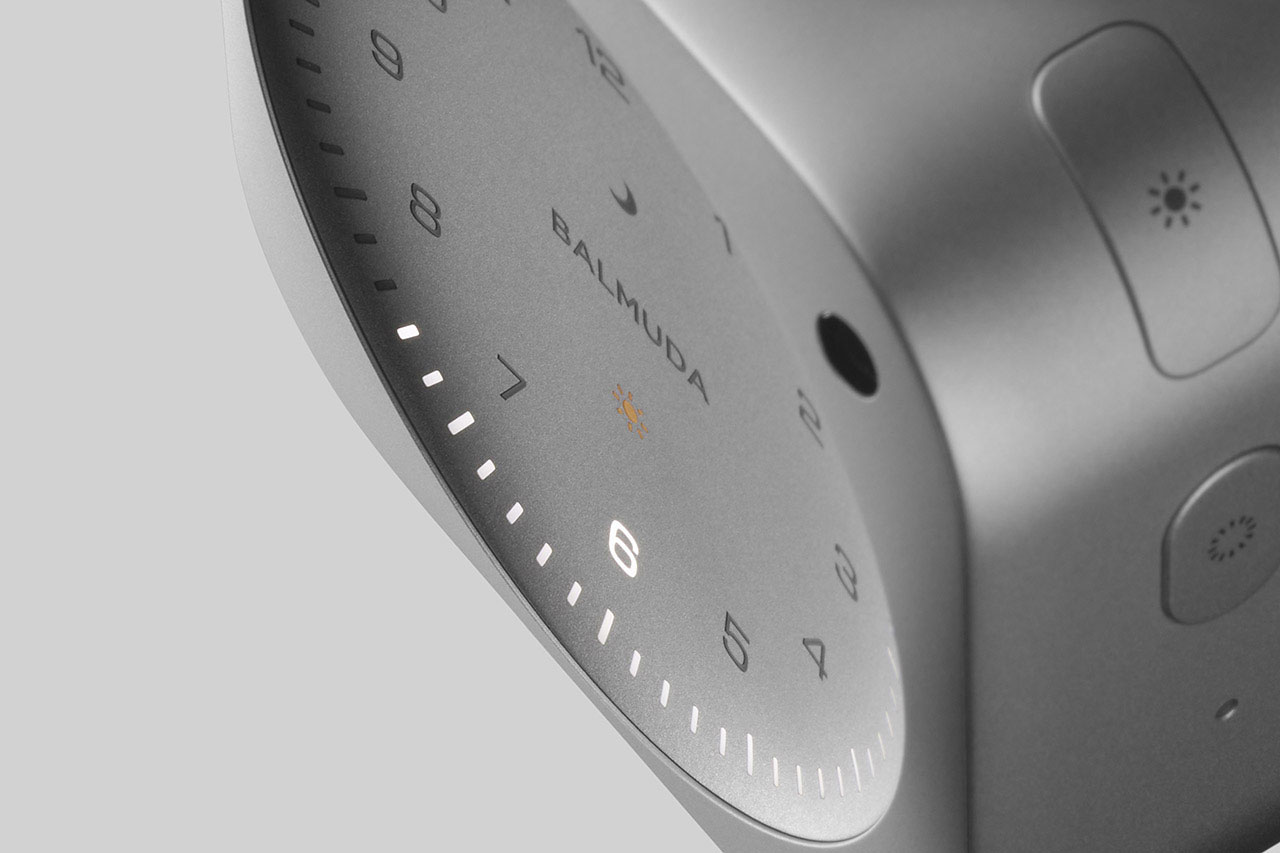 Close-up of a BALMUDA clock displaying a minimalist circular face with numbers, a moon icon, and control buttons on the side.