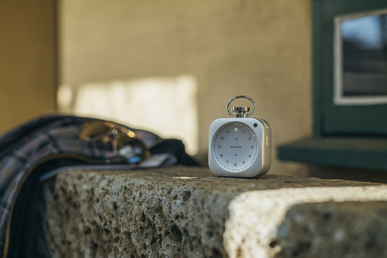 A small square clock with a ring on top sits on a stone ledge next to a plaid jacket and sunglasses, with a blurred window in the background.