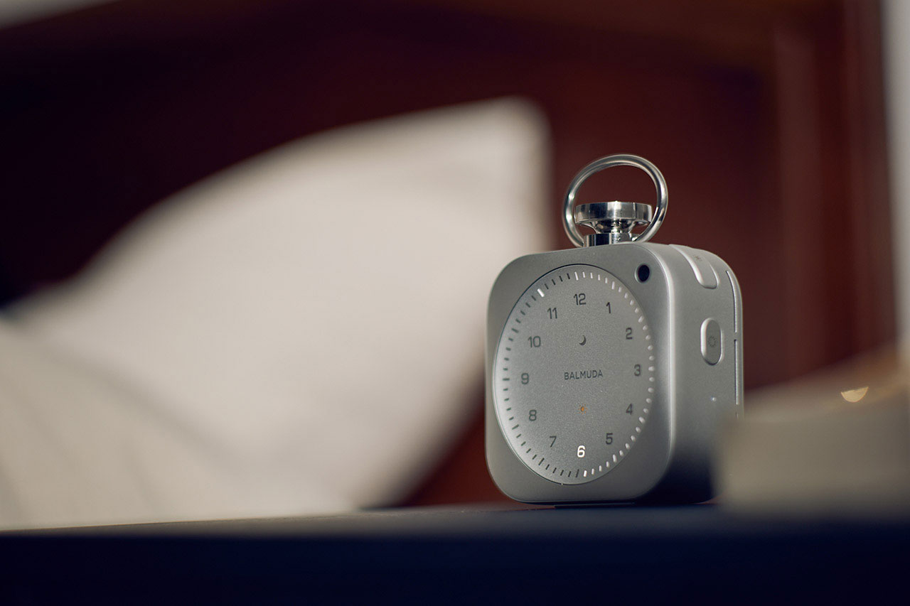 A silver analog alarm clock sits on a bedside table next to a bed with a white pillow and a wooden headboard in the background.