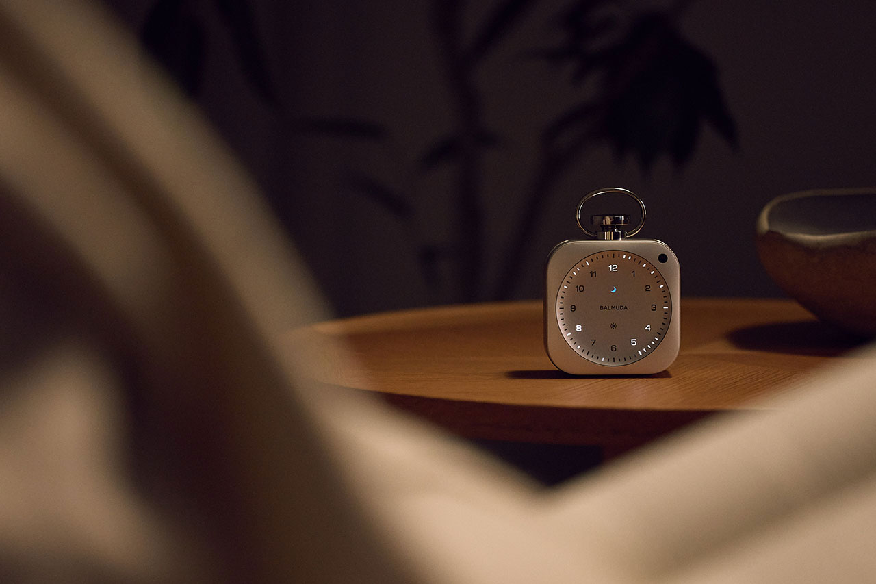 A small, modern analog alarm clock sits on a wooden surface in a dimly lit room, with blurred foreground elements.