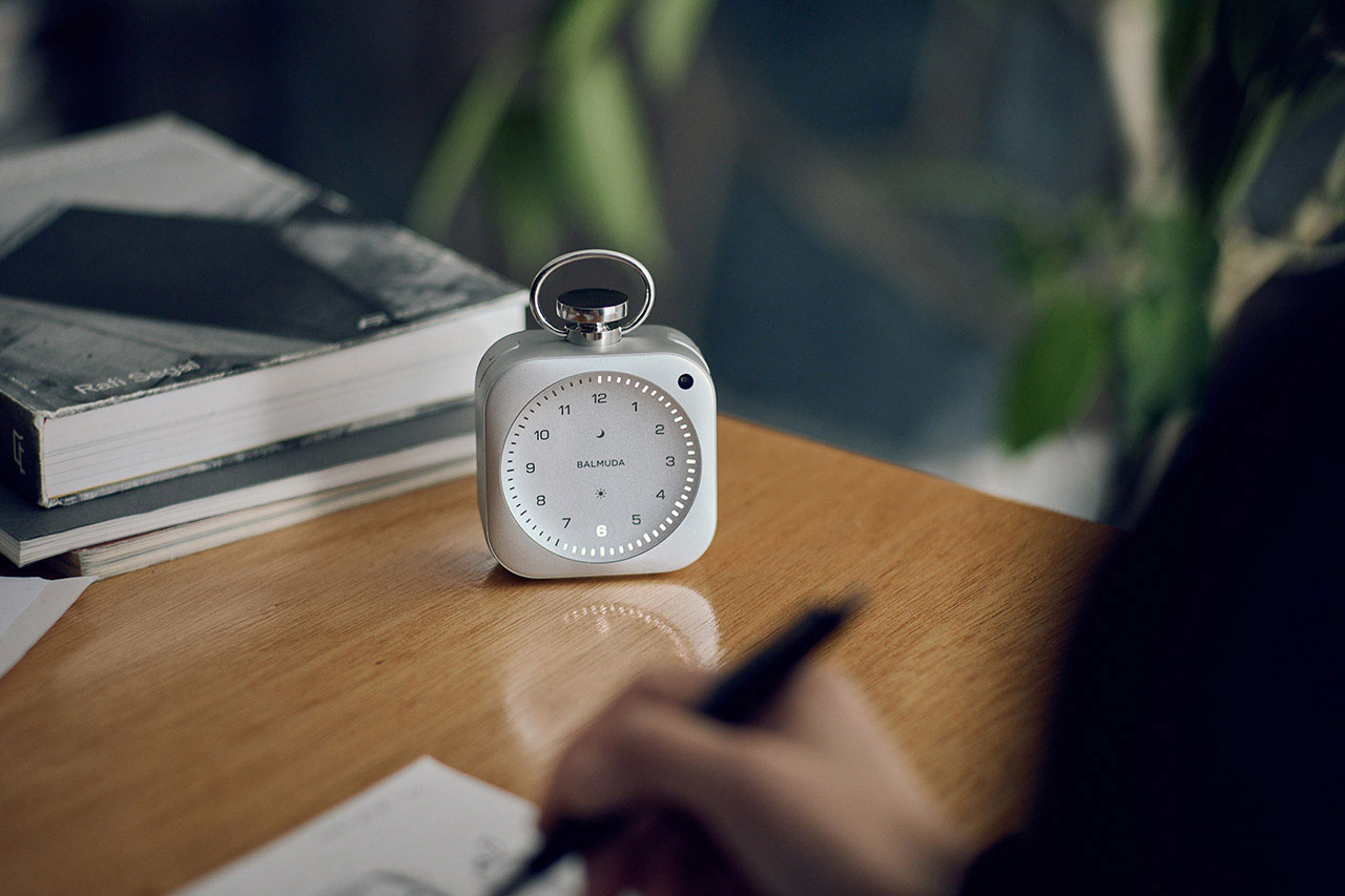 A white analog clock sits on a wooden desk next to a stack of books, with a person writing in the foreground and a blurred green plant in the background.