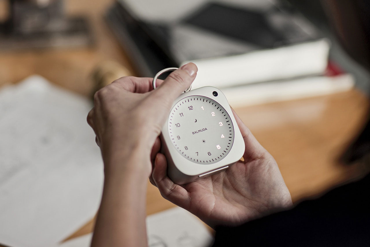 A person holds a small, white analog clock with a circular face and minimalist design, while sitting at a desk with books and papers.