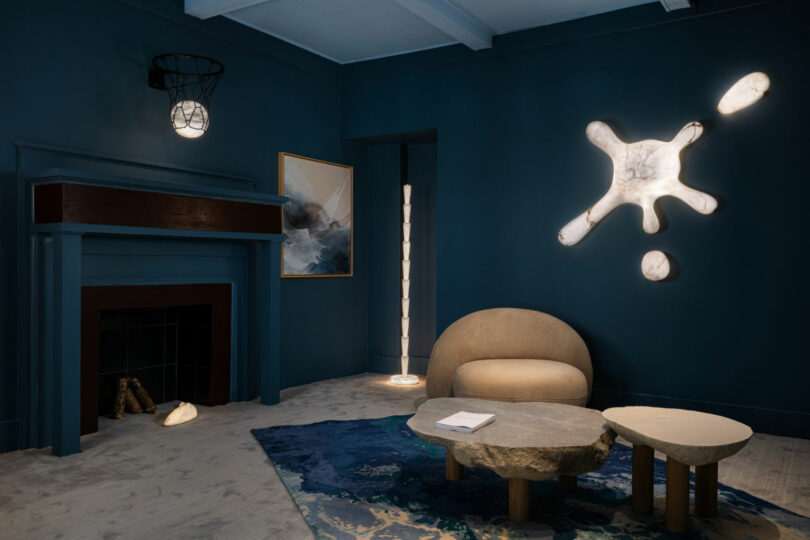 Modern living room with dark blue walls, abstract wall lights, a beige curved sofa, stone coffee tables, and a decorative rug. A basketball hoop light fixture hangs above the fireplace—a perfect Friday Five feature for design inspiration.