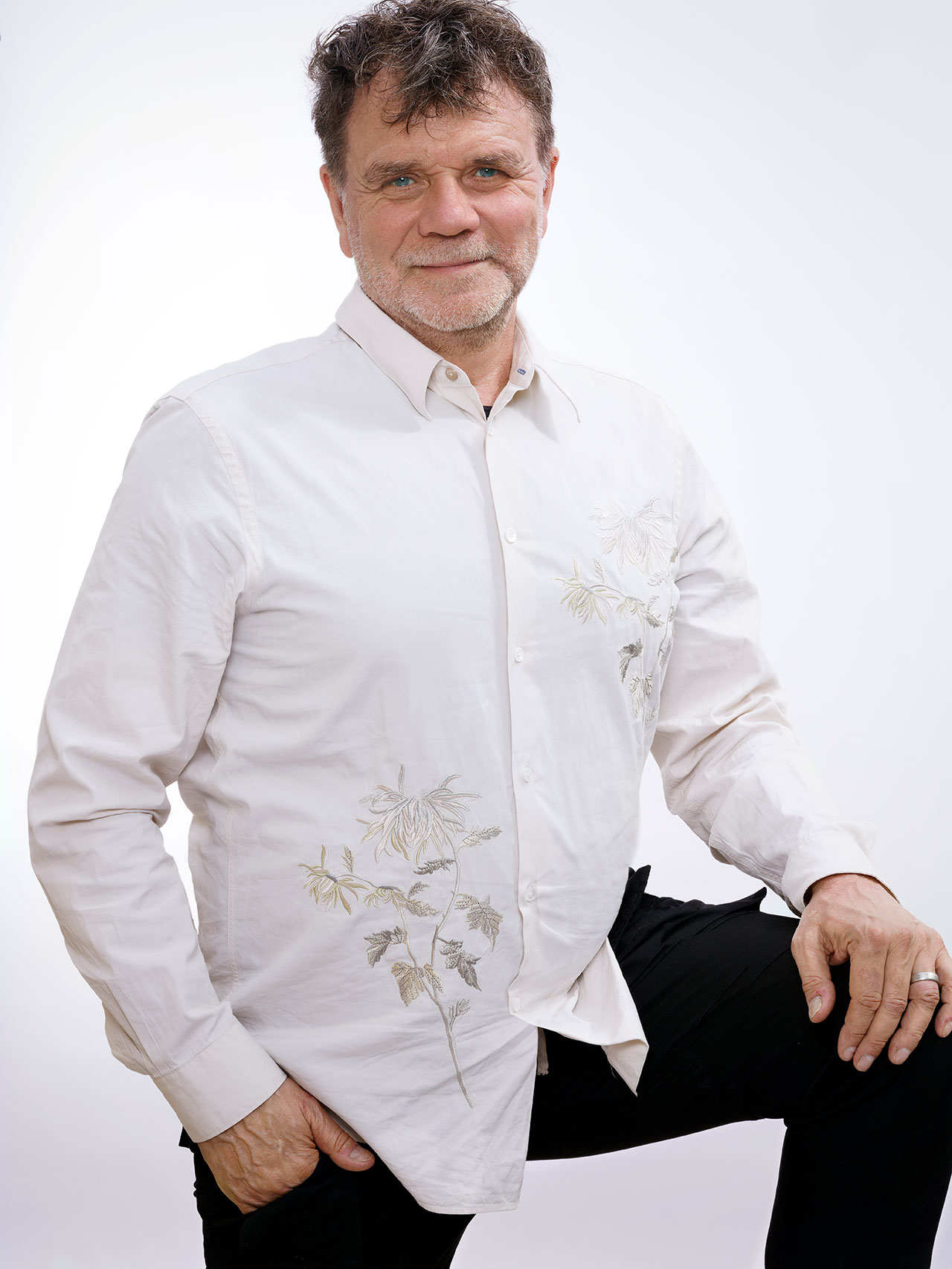 A man with short brown hair and a beard stands with one knee raised, wearing a white Minarc button-up shirt with a floral design and black pants, against a plain white background.