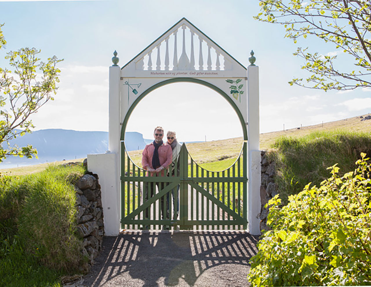 A man and woman stand together behind a decorative green and white gate in a rural outdoor setting with hills and blue sky in the background.